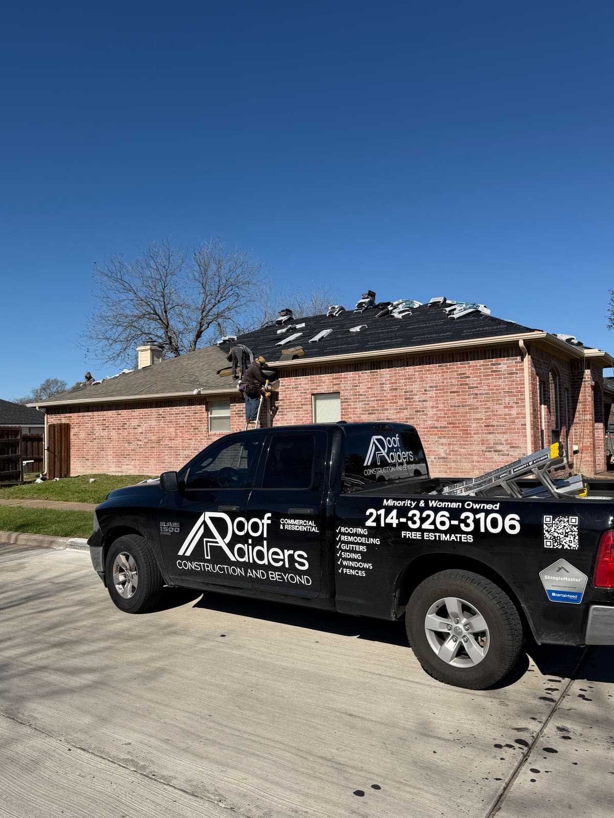 Roof Raiders & Co. truck with the company logo, phone number, Minority and Woman Owned lettering, and CertainTeed ShingleMaster badge parked at an active roof replacement with the crew working on the roof behind it