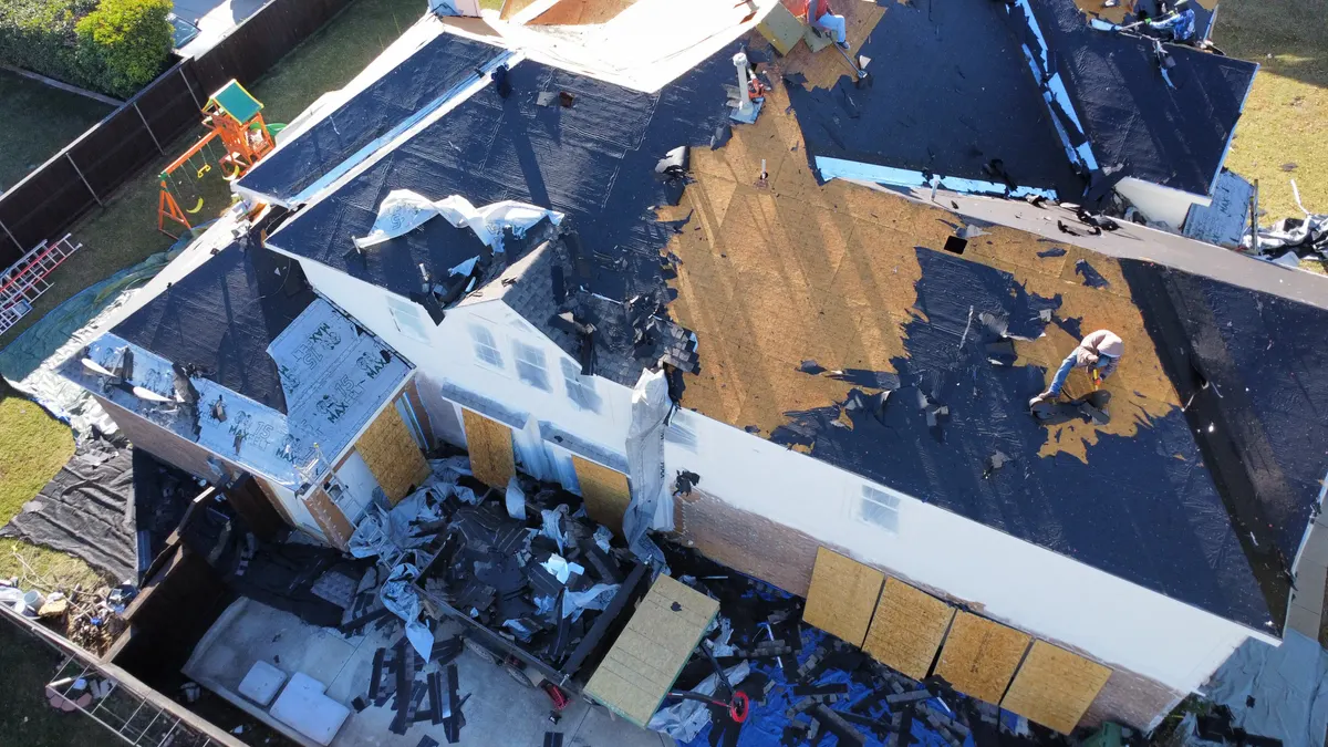 Drone view of a severely storm-damaged two-story North Texas home with boarded windows and exposed roof decking as a Roof Raiders crew works the restoration